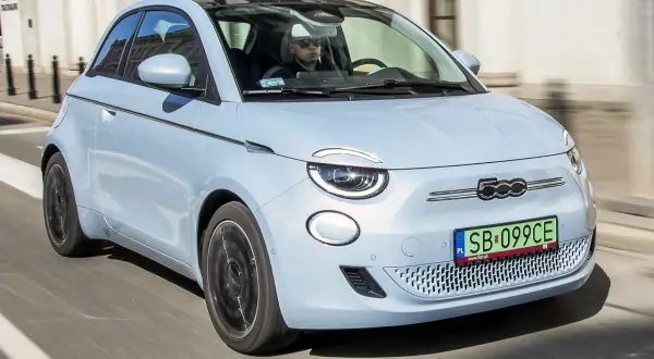 Jasnoniebieski fiat elektryczny 500e z czarnym dachem jedzie ulicą. Kierowca w okularach przeciwsłonecznych prowadzi auto.