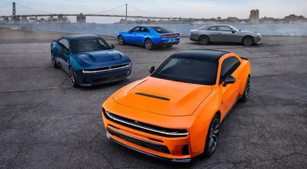 Pomarańczowy i niebieski Dodge Charger Hellcat obok szarego coupe, z mostem i miastem w tle.