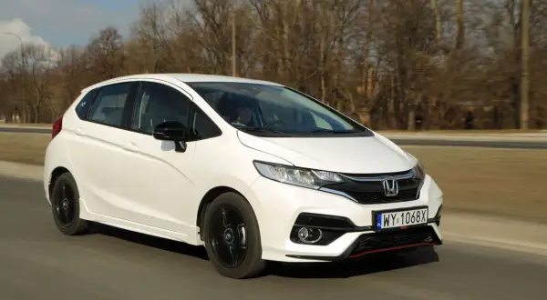 Biały Honda Fit z czarnymi felgami jedzie po drodze, otoczony drzewami.