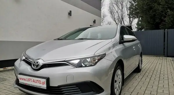 Srebrna Toyota Auris 1.6 benzyna zaparkowana na podjeździe z kostki brukowej, przed nowoczesnym budynkiem.