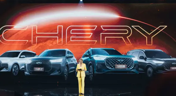 Kobieta w żółtym garniturze prezentuje nowe modele Chery Tiggo. Na ekranie za nią widnieje logo marki.