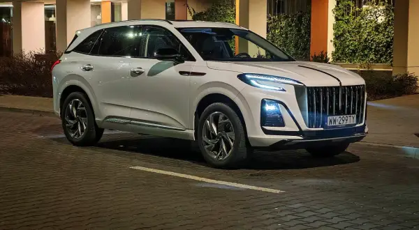 Biały Hongqi HS3 z charakterystycznym grillem i nowoczesnymi światłami stoi na mokrej, brukowanej ulicy nocą.