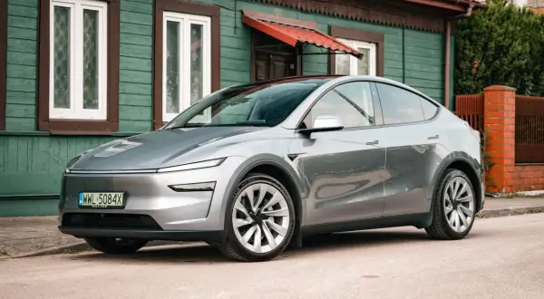 Srebrny Tesla Juniper, nowoczesny samochód elektryczny, zaparkowany przed tradycyjnym, zielonym domem z czerwonym dachem.