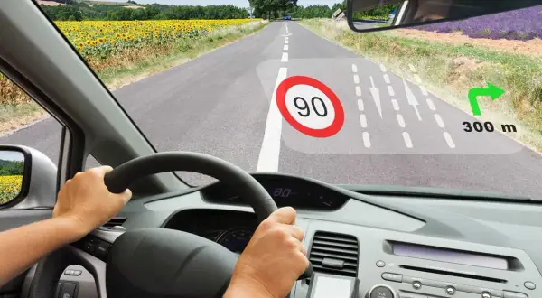 Widok z kabiny samochodu. Na szybie wyświetla się HUD z ograniczeniem prędkości 90, nawigacją i odległością 300 m do skrętu.
