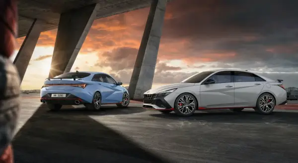 Dwa sportowe samochody Hyundai Elantra N, jeden niebieski, drugi jasnoszary, stoją pod betonowym mostem o zachodzie słońca.