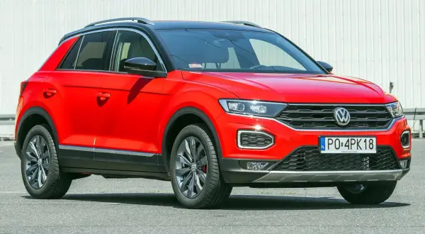 Czerwony, używany Volkswagen T-Roc z czarnym dachem i przyciemnianymi szybami, zaparkowany na tle białej ściany.
