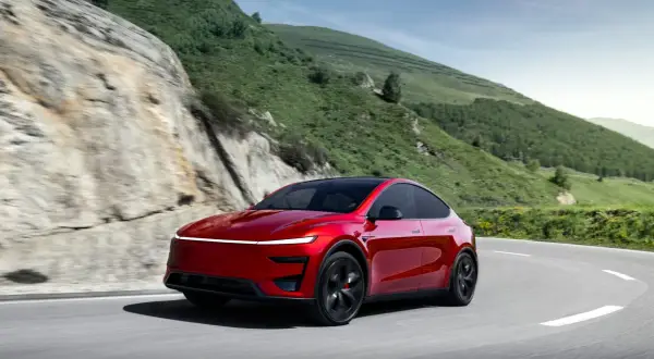 Czerwony Tesla Y Performance pokonuje zakręt na górskiej drodze, otoczony zielonymi wzgórzami i skalistymi zboczami.