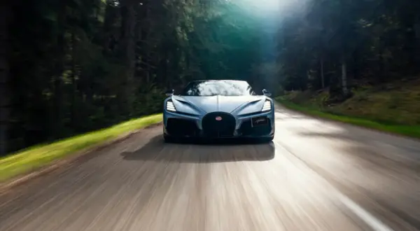 Niebieski Bugatti Mistral pędzi leśną drogą, słońce przebija się przez drzewa.