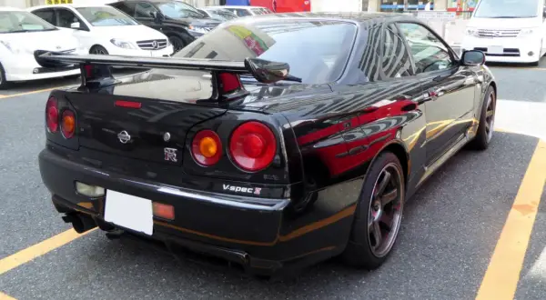 Czarny Nissan GT-R R34 V-spec II z charakterystycznymi okrągłymi światłami i dużym spojlerem, zaparkowany na ulicy.