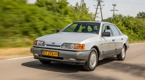 Srebrny Ford Scorpio z lat 90. pędzi po wiejskiej drodze, otoczony zielenią.