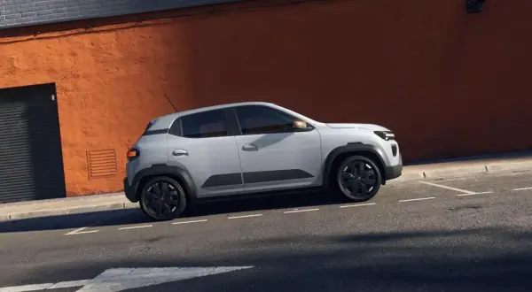 Jasnoszary Dacia Spring na tle pomarańczowej ściany. Sprawdź atrakcyjną dacia spring cena i odkryj jego zalety.