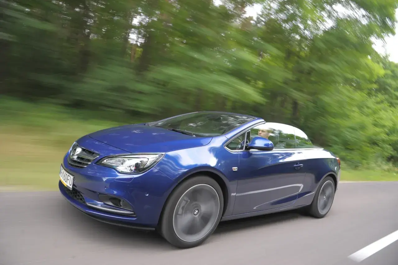 Niebieski Opel Cascada z otwartym dachem pędzi leśną drogą.