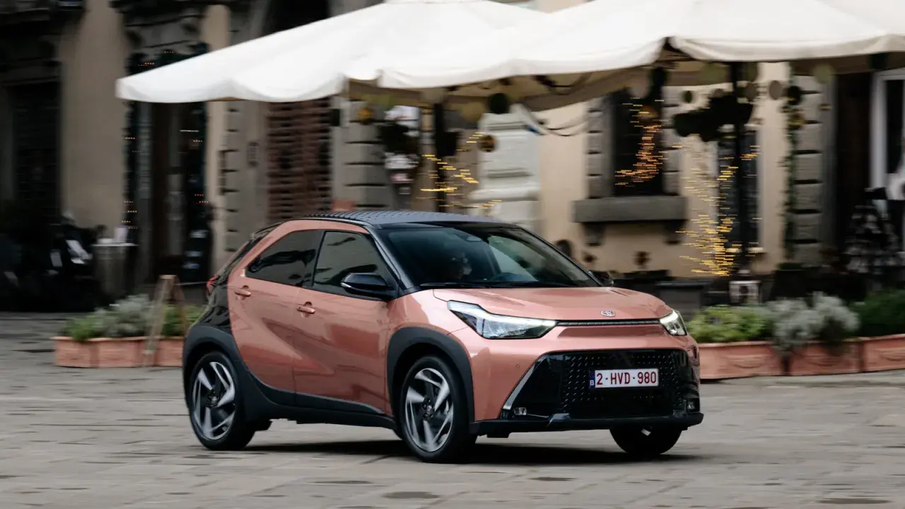 Różowy Toyota Aygo X z czarnym dachem jedzie po brukowanej ulicy.
