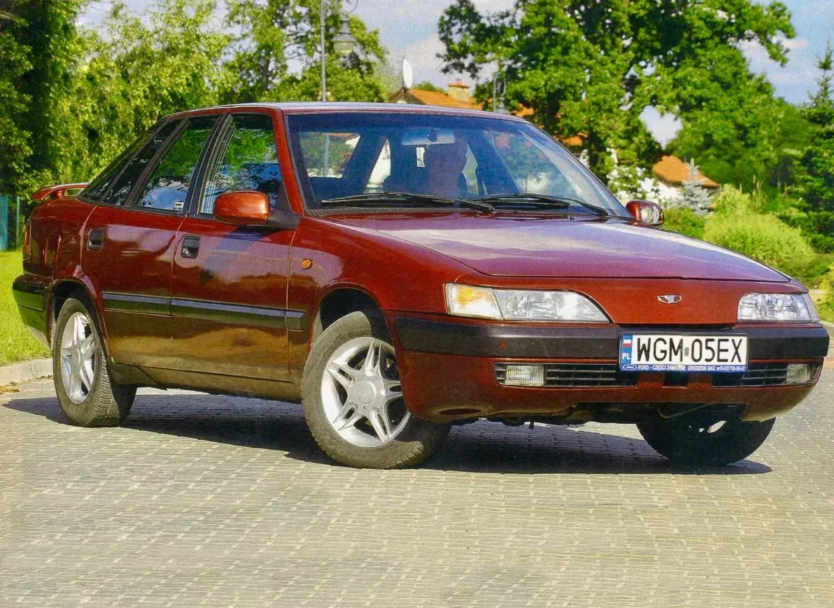 Czerwony Daewoo Espero z lat 90. stoi na brukowanej drodze, otoczony zielenią.