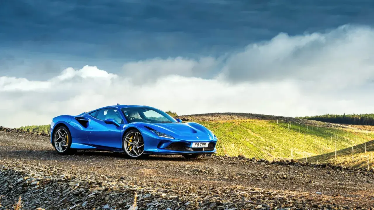 Niebieskie Ferrari F8 na tle malowniczego, pagórkowatego krajobrazu z chmurami na niebie.