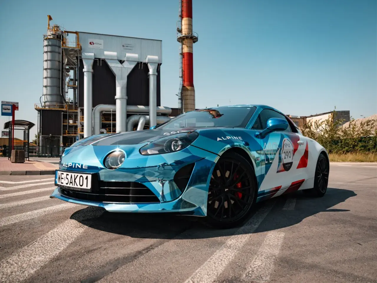 Błękitno-białe Alpine A110S z numerem 290 na tle przemysłowej zabudowy.