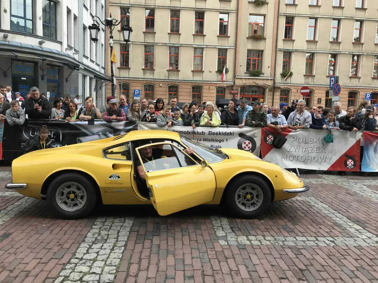 Żółte Ferrari Dino z otwartymi drzwiami, kierowca w środku, tłum widzów za barierką.