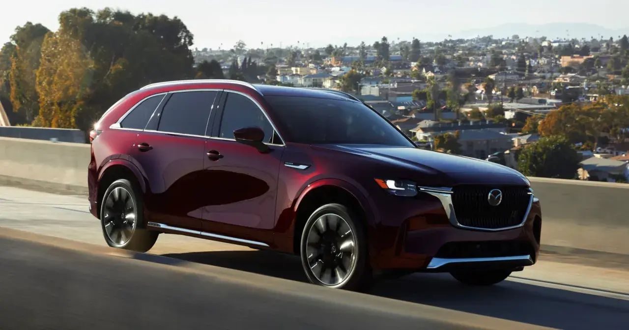 Elegancka, bordowa Mazda CX-90 sunie po drodze na tle malowniczego, miejskiego krajobrazu.