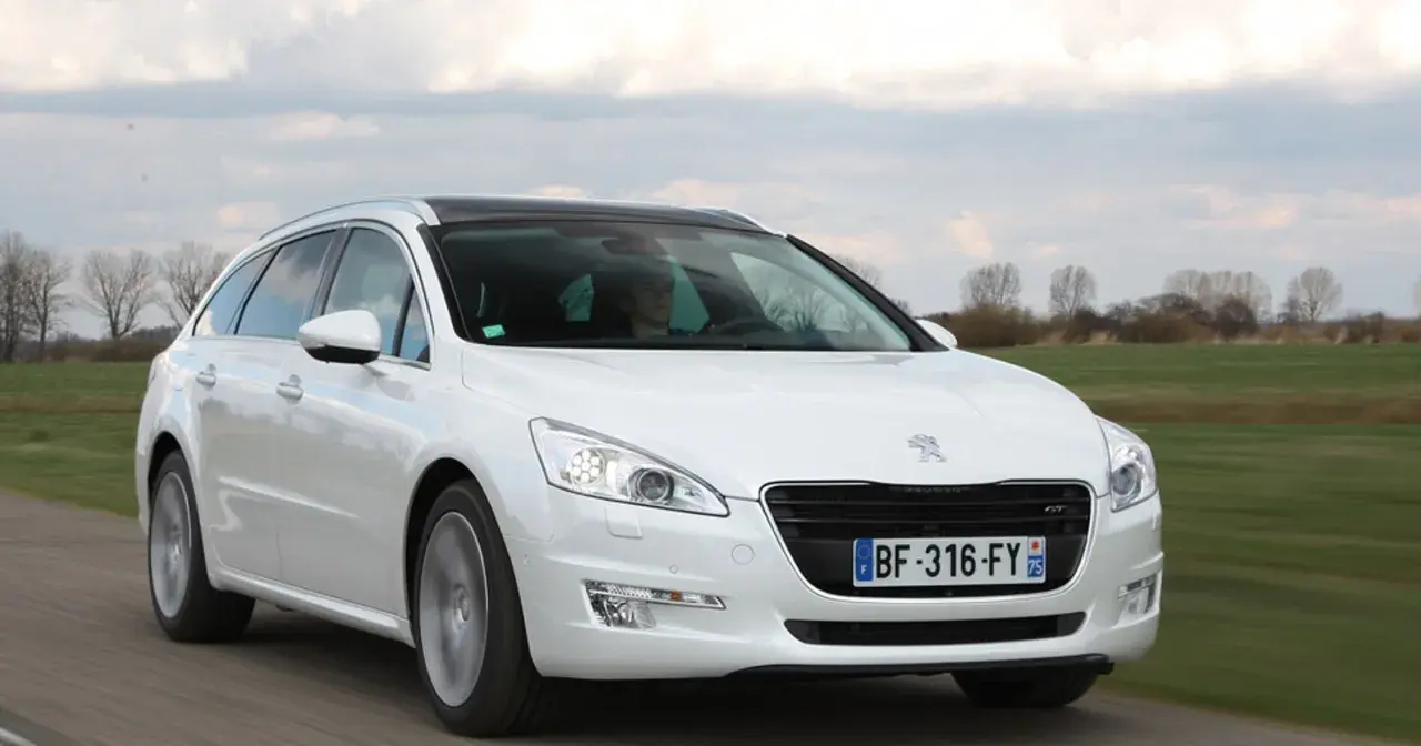 Biały Peugeot 508 SW z czarnym dachem jedzie po drodze, otoczony zielonymi polami i drzewami.