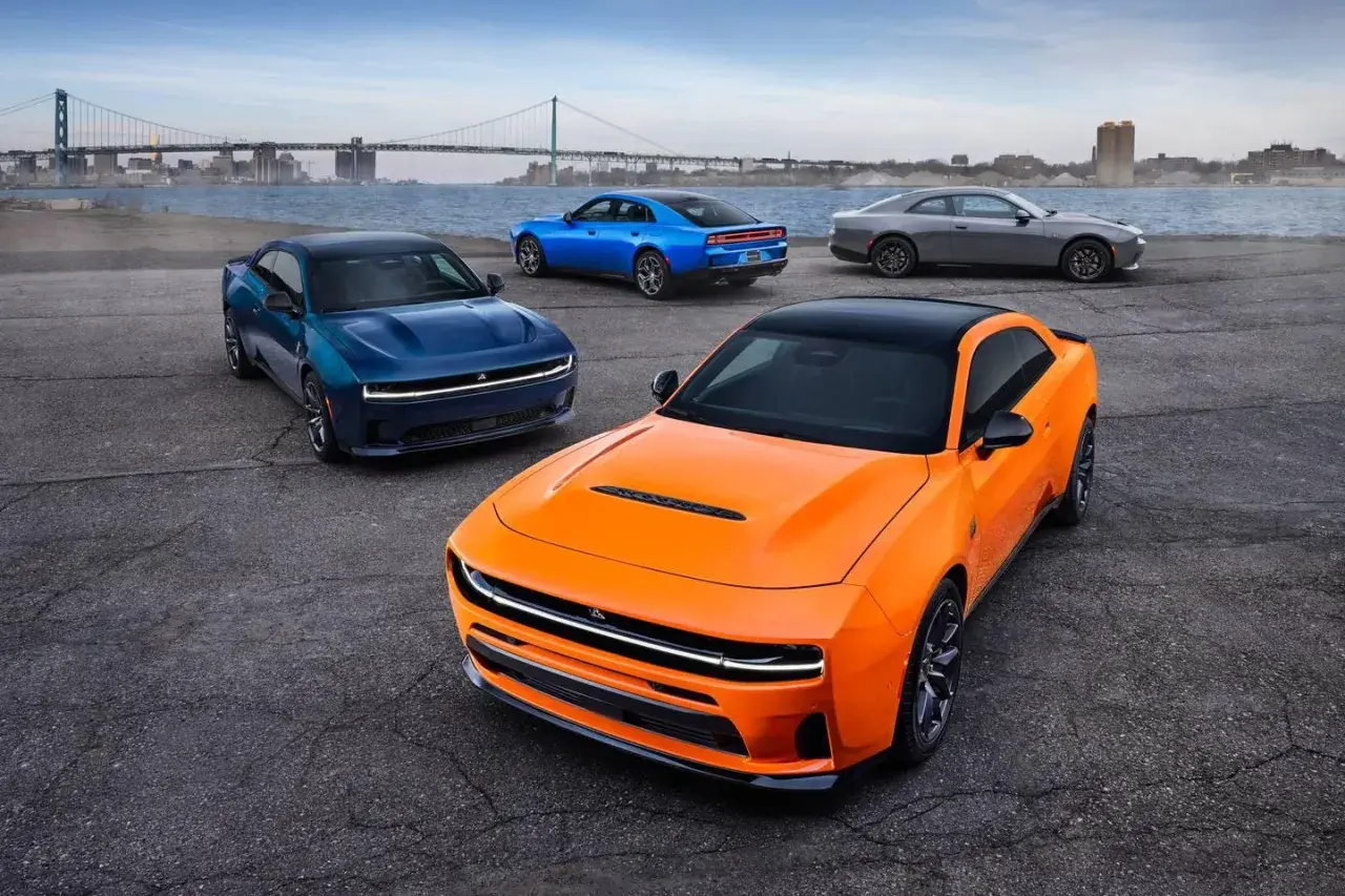 Pomarańczowy i niebieski Dodge Charger Hellcat obok szarego coupe, z mostem i miastem w tle.