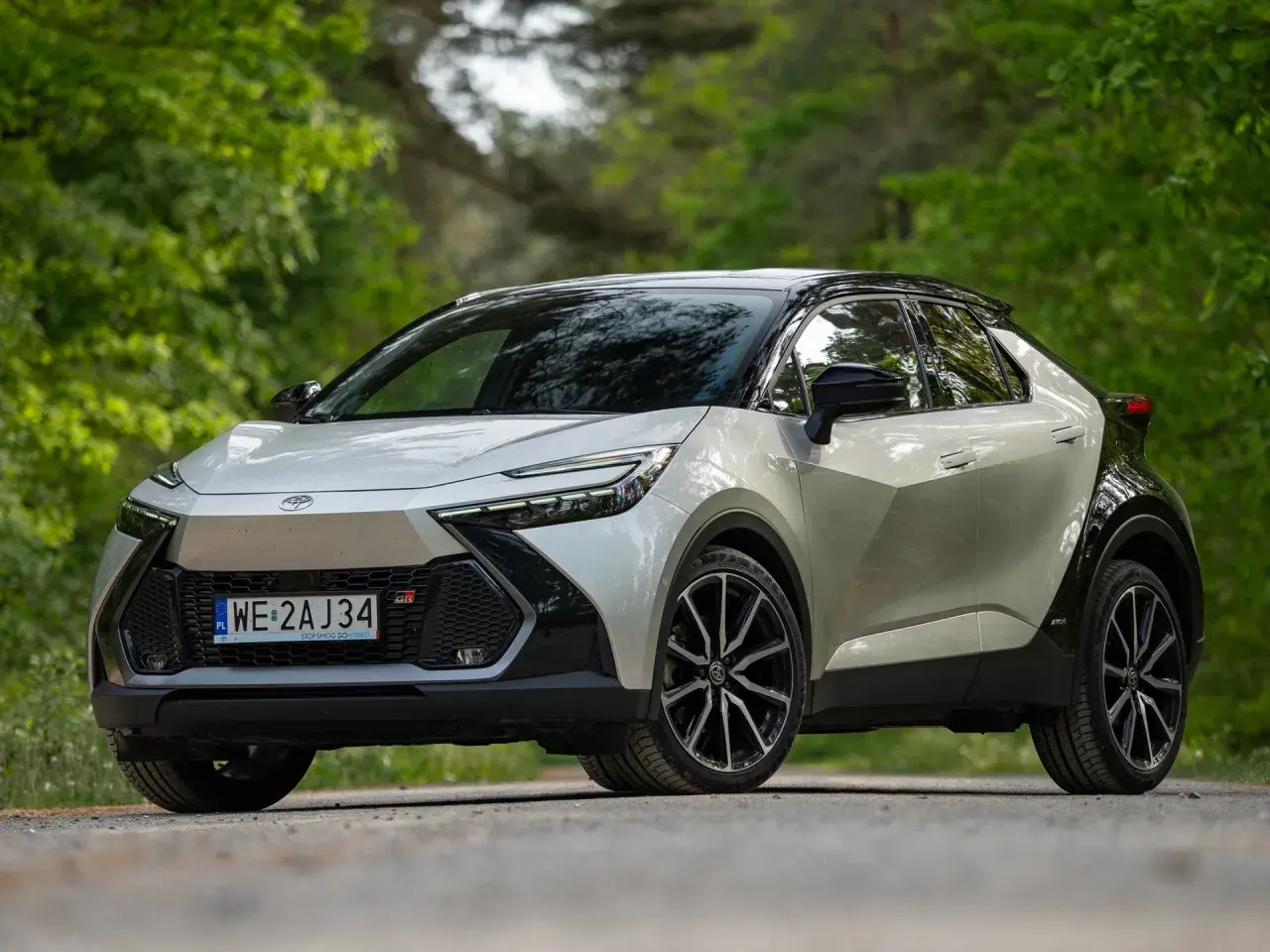 Nowy Toyota C-HR GR Sport w kolorze srebrnym z czarnym dachem, zaparkowany na leśnej drodze. Sprawdź atrakcyjną toyota chr cena!