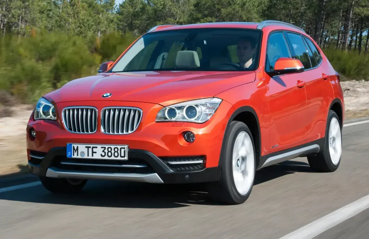 Pomarańczowy BMW X1 E84 w ruchu, z kierowcą za kierownicą, na tle drzew.