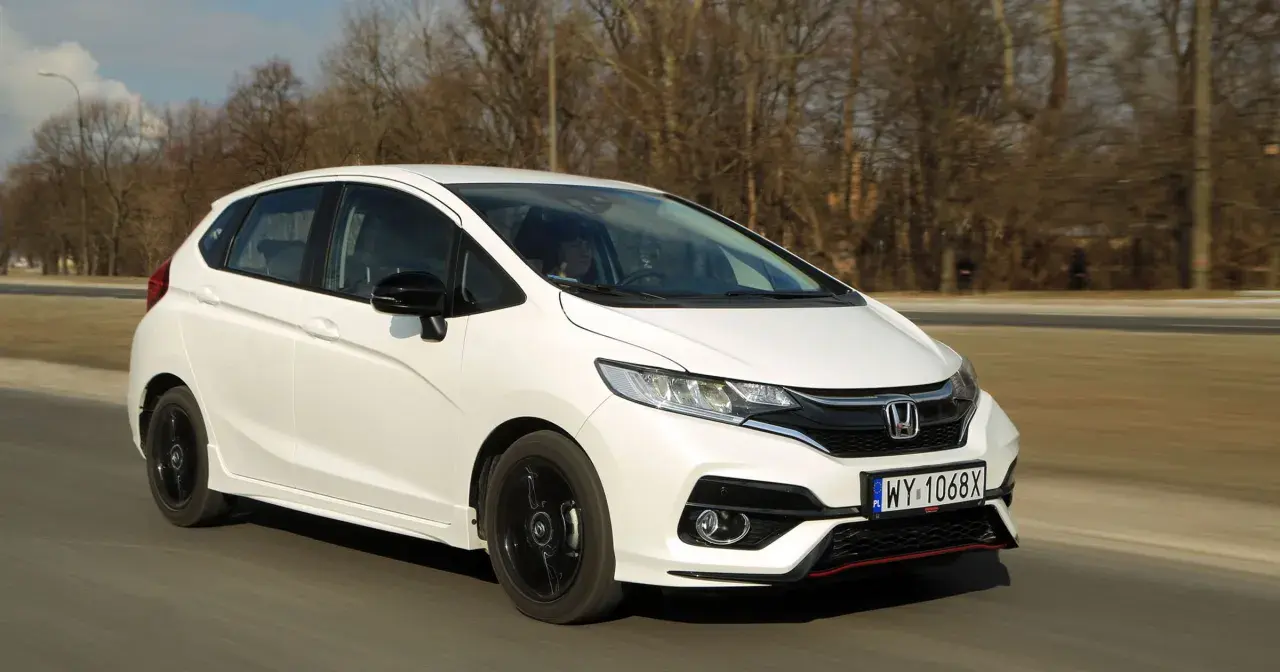 Biały Honda Fit z czarnymi felgami jedzie po drodze, otoczony drzewami.