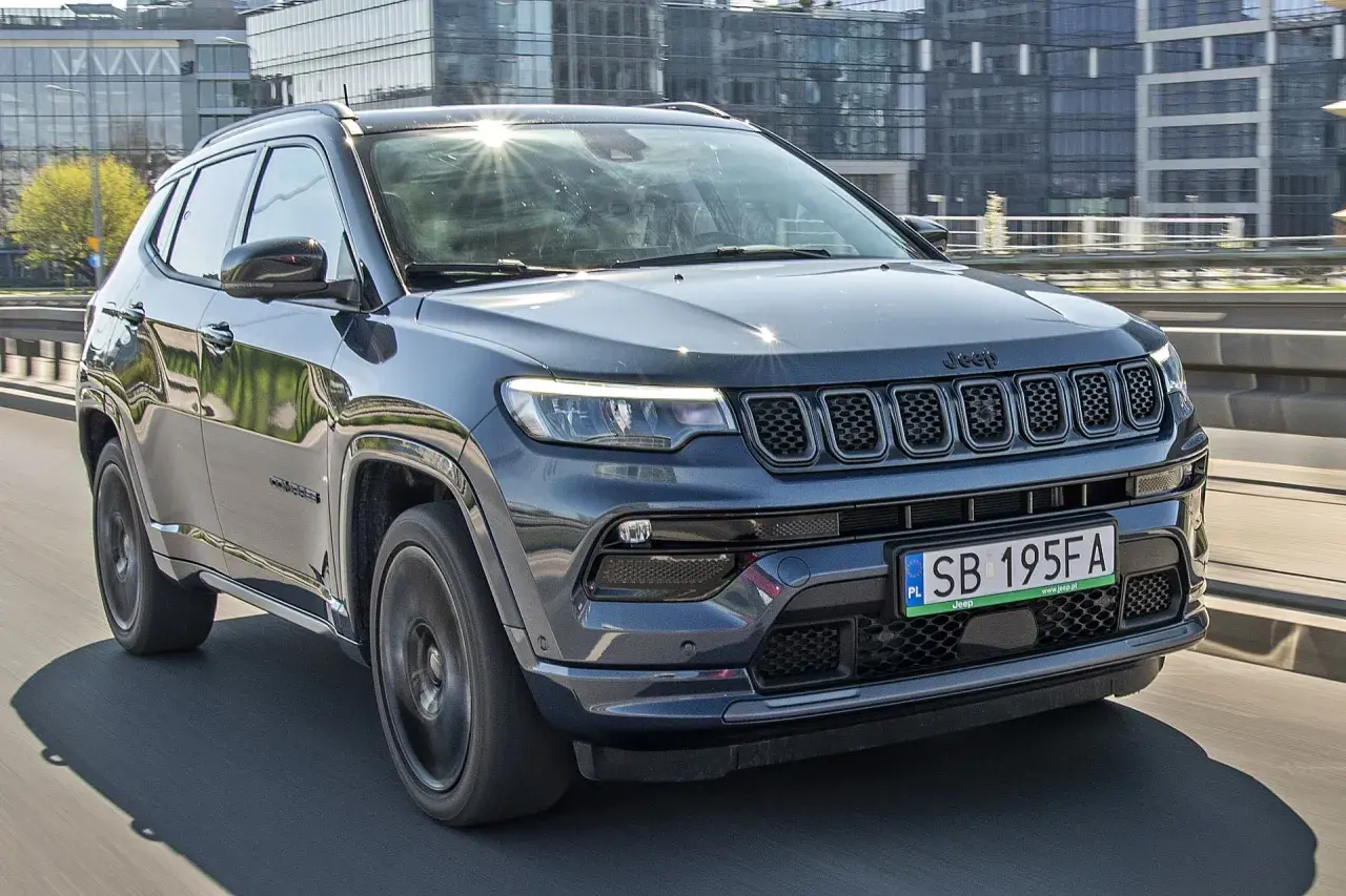 Ciemnoszary Jeep Compass 2025 na drodze, z nowoczesnymi budynkami w tle.