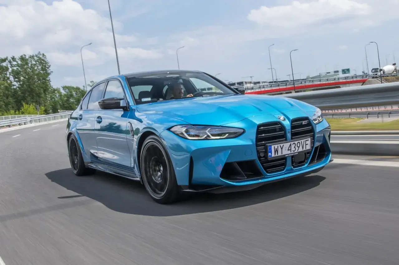 Niebieskie BMW M3 Competition pędzi po drodze, błyszcząc w słońcu.