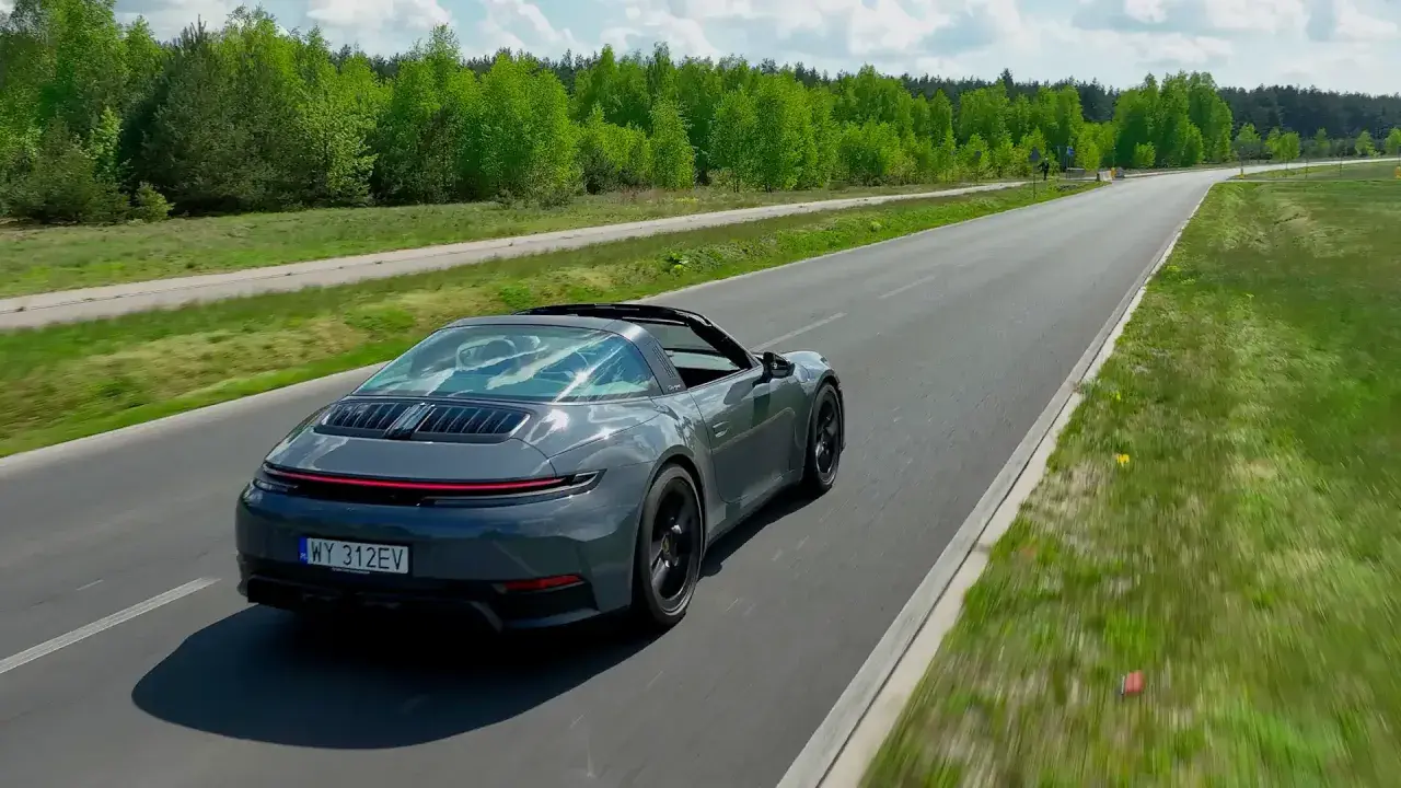 Szary Porsche Targa sunie po asfaltowej drodze, otoczony zielenią.