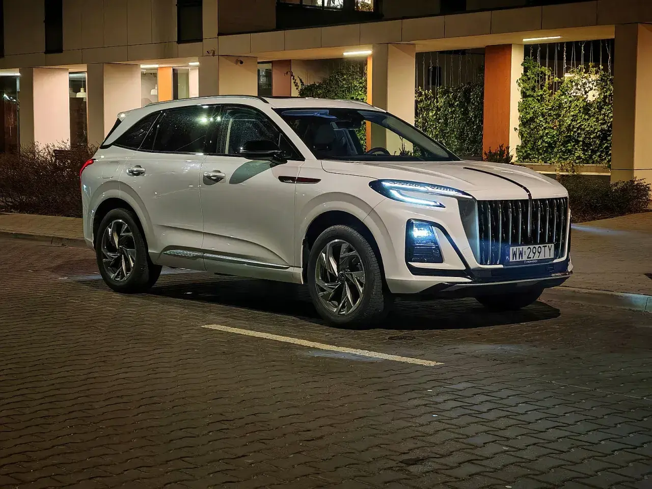 Biały Hongqi HS3 z charakterystycznym grillem i nowoczesnymi światłami stoi na mokrej, brukowanej ulicy nocą.