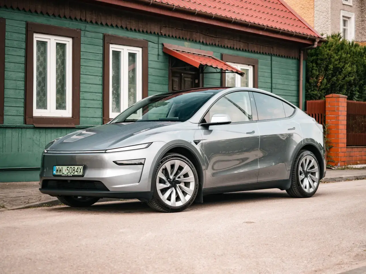 Srebrny Tesla Juniper, nowoczesny samochód elektryczny, zaparkowany przed tradycyjnym, zielonym domem z czerwonym dachem.