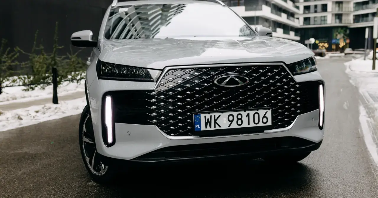 Biały Tiggo 4 z charakterystycznym grillem i pionowymi światłami LED, zaparkowany na mokrej drodze.