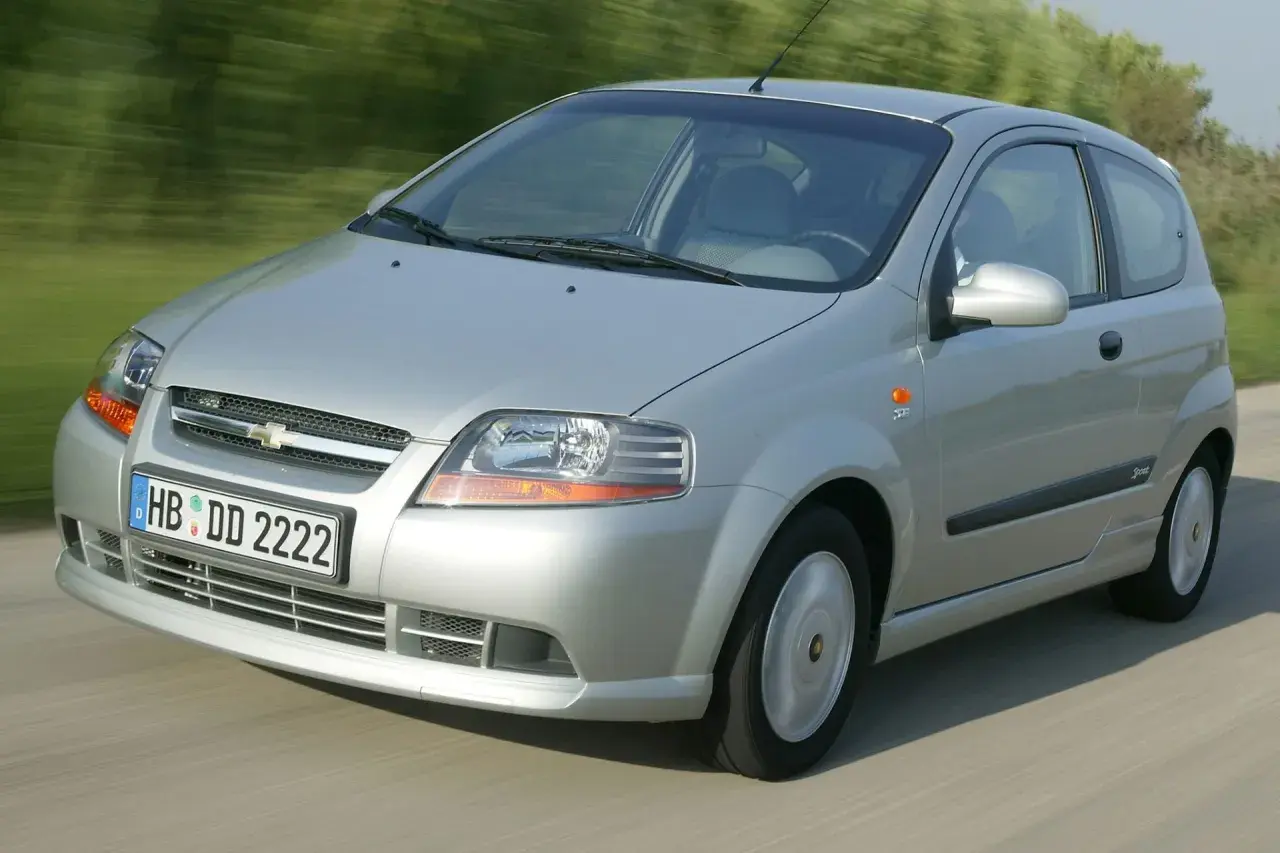 Srebrny Chevrolet Kalos w wersji 3-drzwiowej jedzie po drodze, otoczony zielenią.