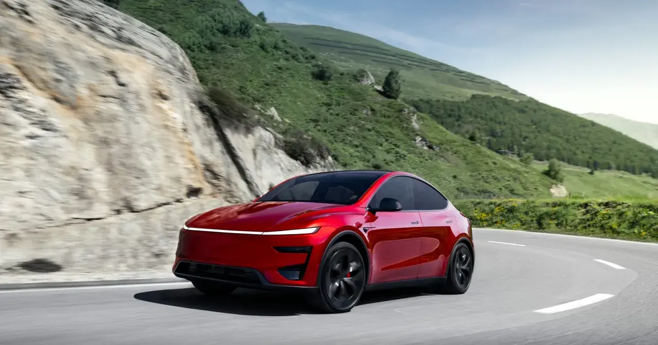 Czerwony Tesla Y Performance pokonuje zakręt na górskiej drodze, otoczony zielonymi wzgórzami i skalistymi zboczami.