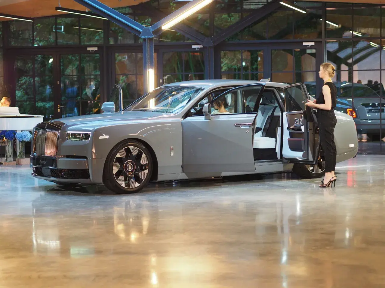 Elegancki szary Rolls-Royce Phantom z otwartymi drzwiami, obok stoi kobieta. Poznaj jego imponującą rolls royce cena.