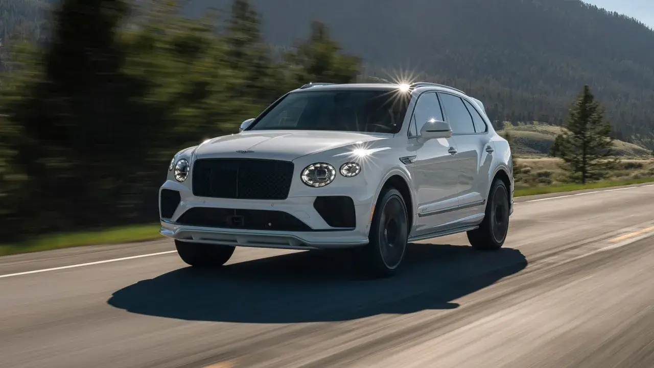 Biały Bentley SUV pędzi po krętej drodze wśród zielonych wzgórz.