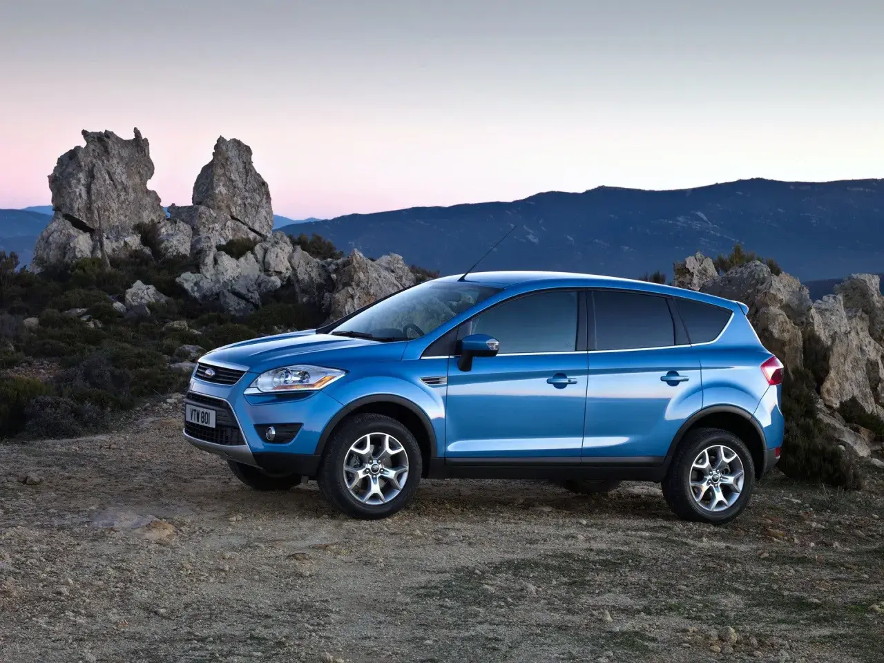 Niebieski Ford Kuga MK1 na tle skalistego krajobrazu o zachodzie słońca.