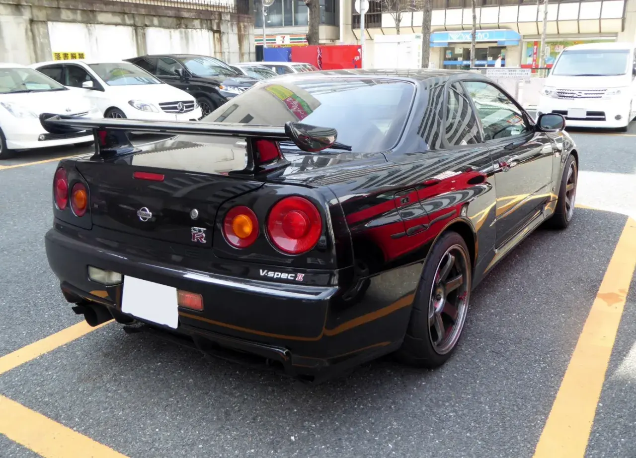Czarny Nissan GT-R R34 V-spec II z charakterystycznymi okrągłymi światłami i dużym spojlerem, zaparkowany na ulicy.