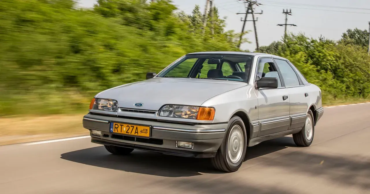 Srebrny Ford Scorpio z lat 90. pędzi po wiejskiej drodze, otoczony zielenią.