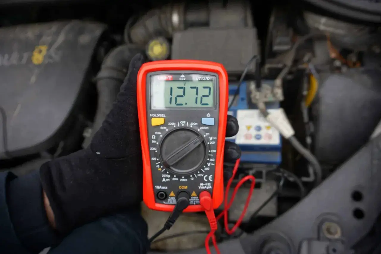 Ręka w czarnej rękawiczce trzyma multimetr pokazujący 12.72V, co jest kluczowe, jak sprawdzić akumulator.