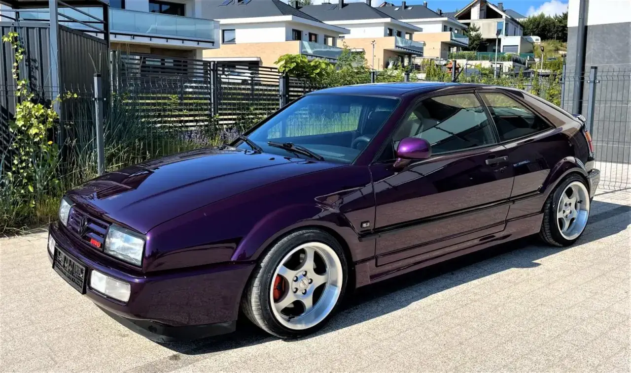 Lśniący, fioletowy Volkswagen Corrado VR6 prezentuje się okazale na tle nowoczesnych domów.
