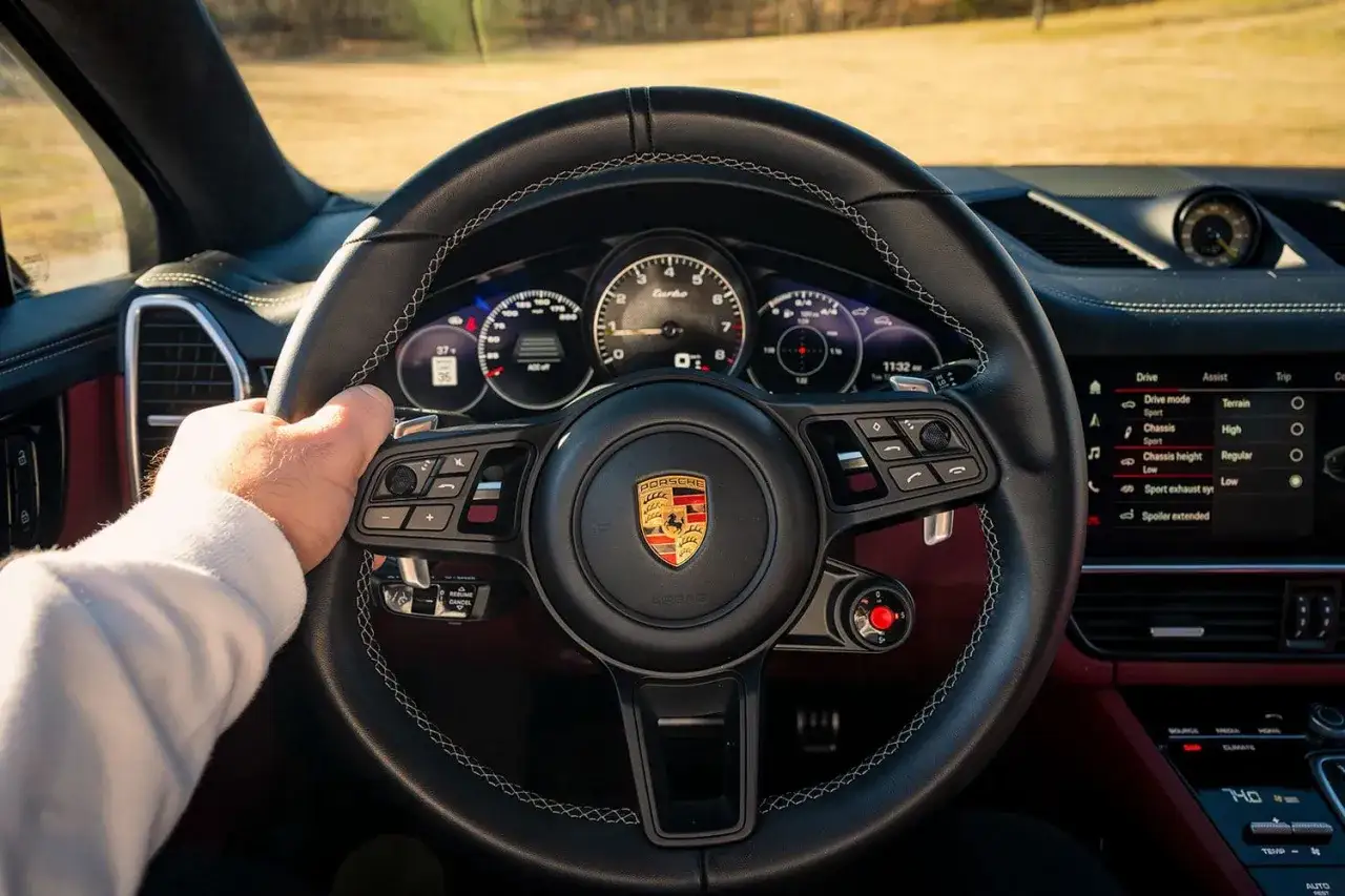 Ręka na kierownicy Porsche Cayenne Turbo GT. Widok na deskę rozdzielczą z ekranem multimediów i zegarami.