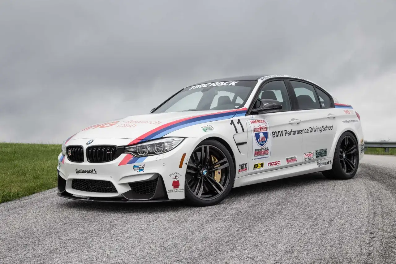 Białe BMW M3 Competition z numerem 11 i licznymi sponsorami, gotowe do jazdy na torze.