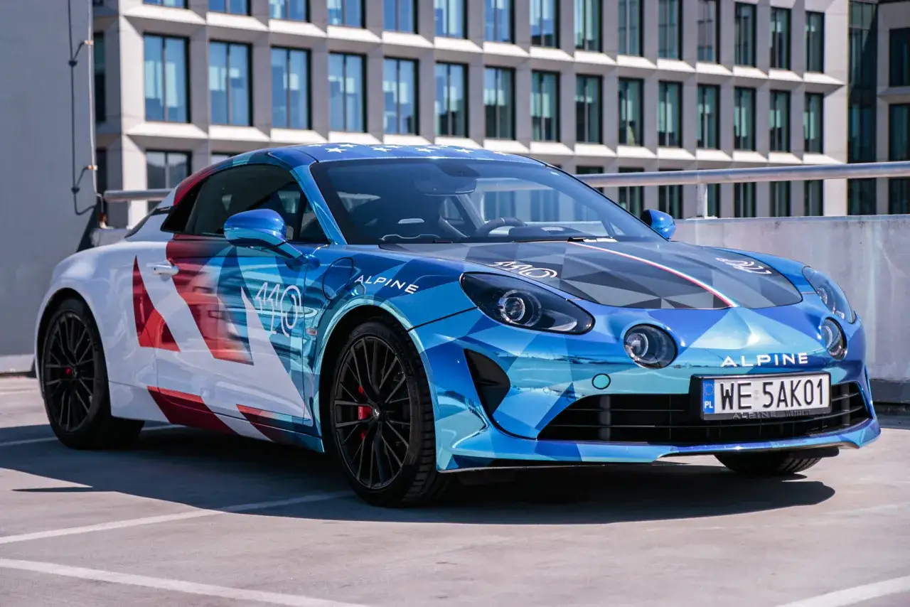 Błękitny Alpine A110S z grafiką w kształcie rombów i białymi akcentami, zaparkowany przed nowoczesnym budynkiem.