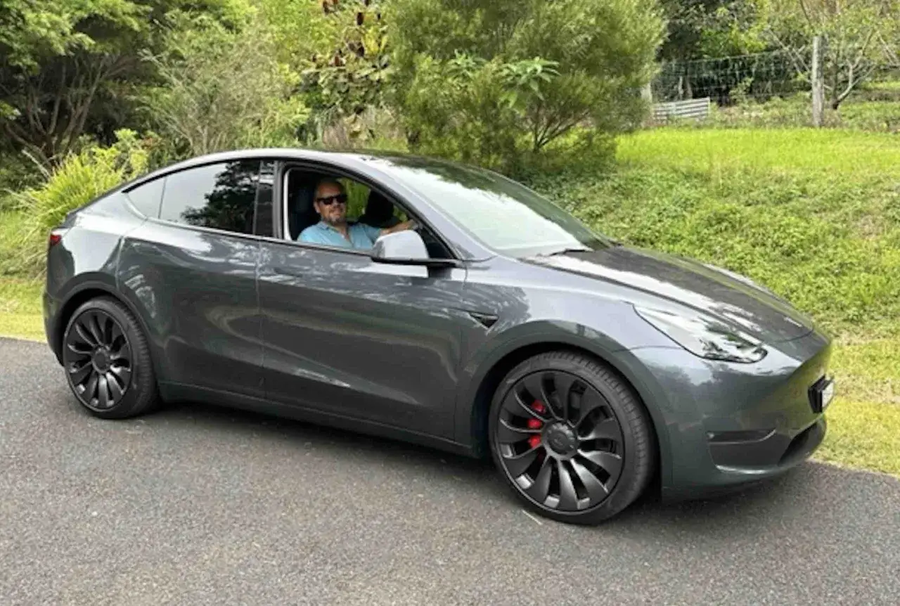 Mężczyzna w okularach przeciwsłonecznych siedzi za kierownicą swojego ciemnoszarego Tesla Y Performance, gotowy na przejażdżkę po malowniczej okolicy.