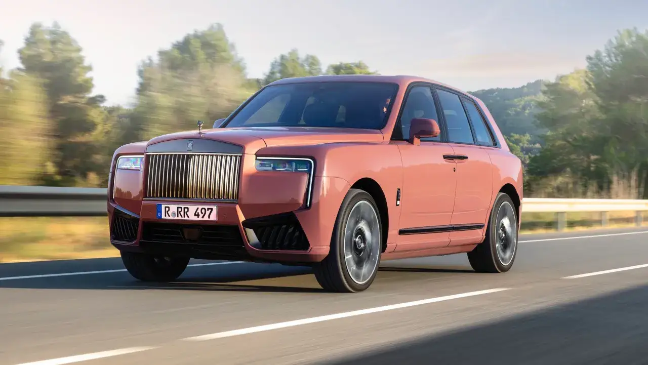 Różowy Rolls-Royce Cullinan sunie po drodze, otoczony zielenią.
