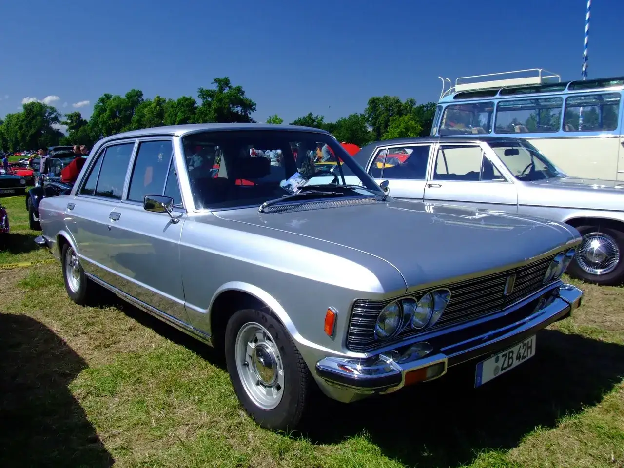 Srebrny fiat 130 z lat 70. prezentuje się elegancko na trawie, obok innych klasyków.