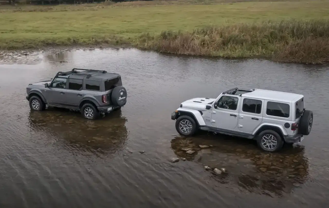 Szary Ford Bronco i biały Jeep Wrangler Rubicon stoją w płytkiej wodzie, gotowe na przygodę.