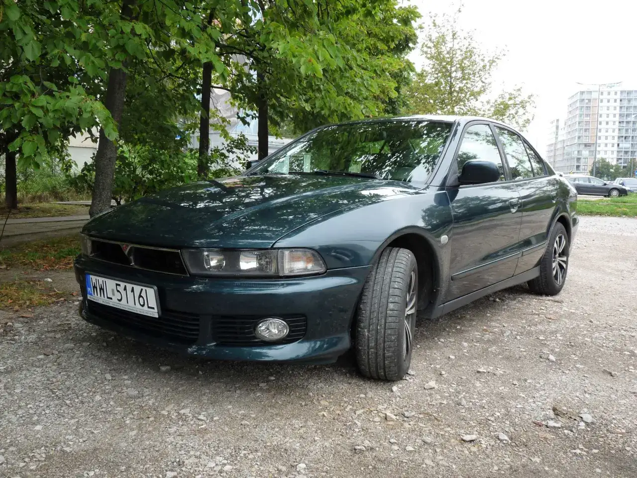 Zielony Mitsubishi Galant, klasyk z lat 90., zaparkowany na szutrowej drodze, otoczony zielenią.